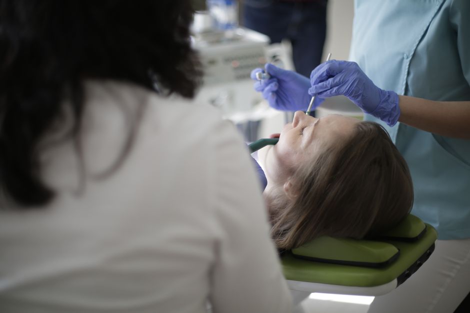Crop anonymous female dentist in blue uniform and latex gloves using drill and mouth mirror while treating teeth of female patient lying in dental chair with opened mouth and closed eyes
